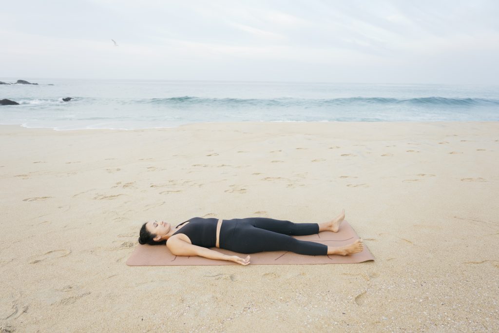 Young woman wearing sportswear practicing yoga savasana pose relaxing on mat at the beach with ocean waves in the background enjoying a mindful moment. healthy lifestyle concept