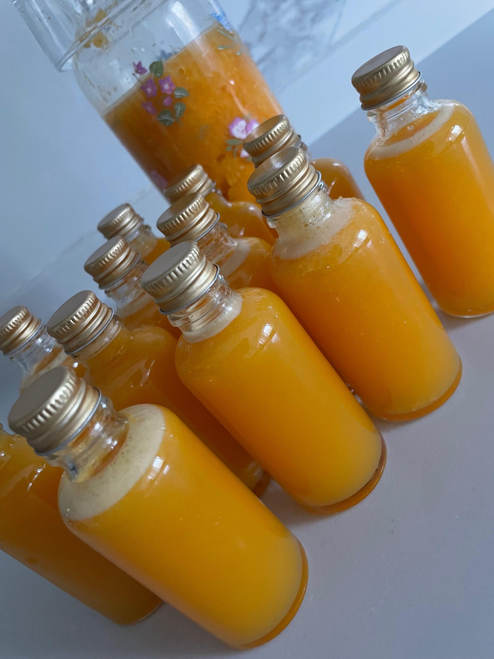 Small glass bottles filled with orange liquid are arranged on a table, with a larger container of the same liquid in the background—perfect for cozymoodallday.com’s article about ginger shots for health and skin care.