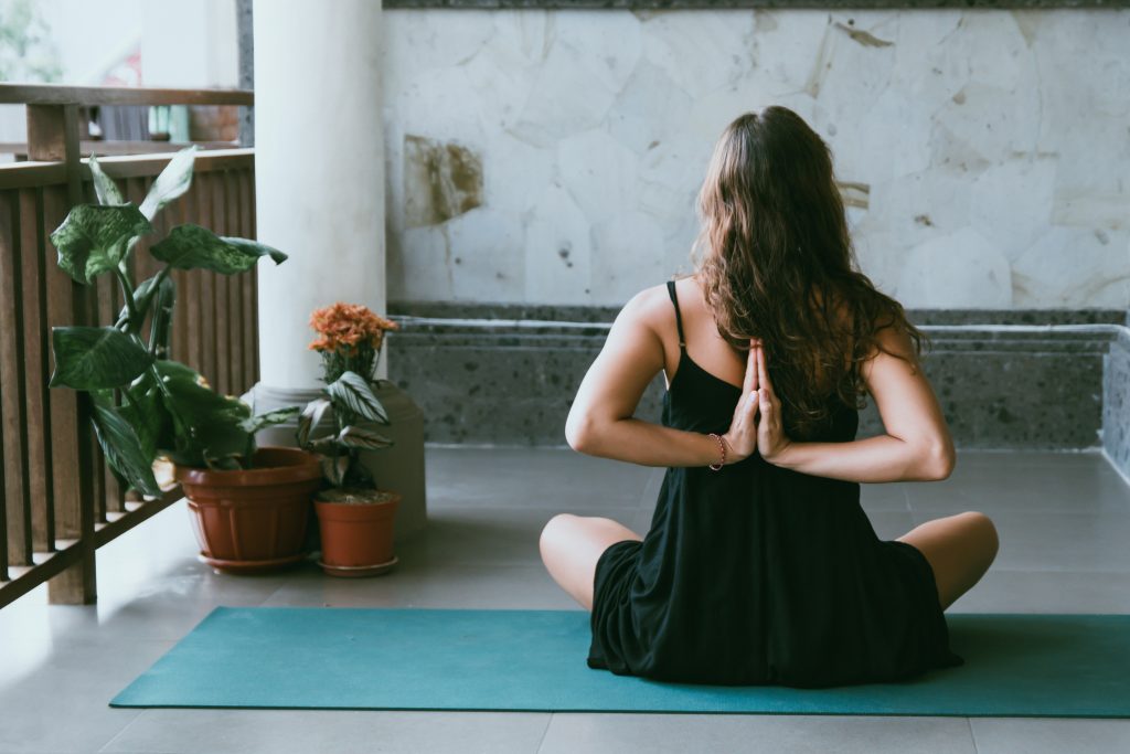 A person sits cross-legged on a yoga mat indoors, performing a reverse prayer pose with hands behind their back. Potted plants are placed nearby, inspiring you to build your own yoga space at home.