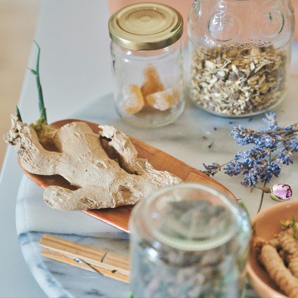 A marble tray for cozymoodallday.com holds dried ginger root, glass jars with dried herbs and sugar cubes, a sprig of lavender, and wooden clothespins—perfect props for an article about ginger shots for health and skin care.