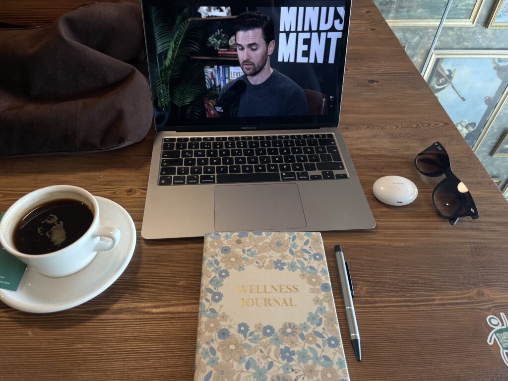 A minimal and cozy workspace featuring a cup of coffee, a wellness journal, a pen, and a MacBook Air on a wooden table. The laptop screen displays a calming coastal landscape, while natural light creates a warm and peaceful atmosphere for learning about digital detox benefits.
