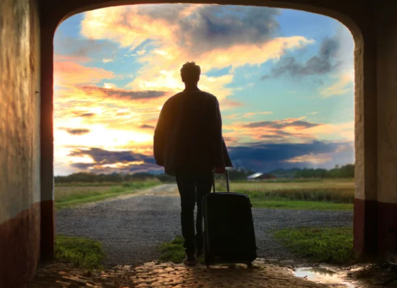 Benefits of Travelling: Why Travelling Is Good for Your Mood Benefits of travelling shown by a solo traveler with luggage walking toward a peaceful sunset countryside road.