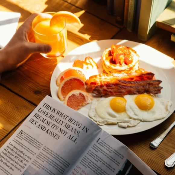 Alt text: High-protein breakfast with fried eggs, crispy bacon, pancakes topped with strawberries, fresh grapefruit slices, and a glass of orange juice on a wooden table.