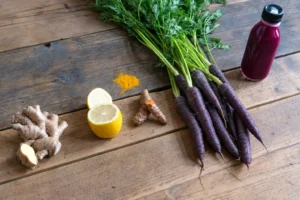 Black Carrot Juice: Easy Homemade Recipe and Benefits Black carrots with leafy green tops beside a bottle of purple juice, lemon, ginger and turmeric on a wooden table.