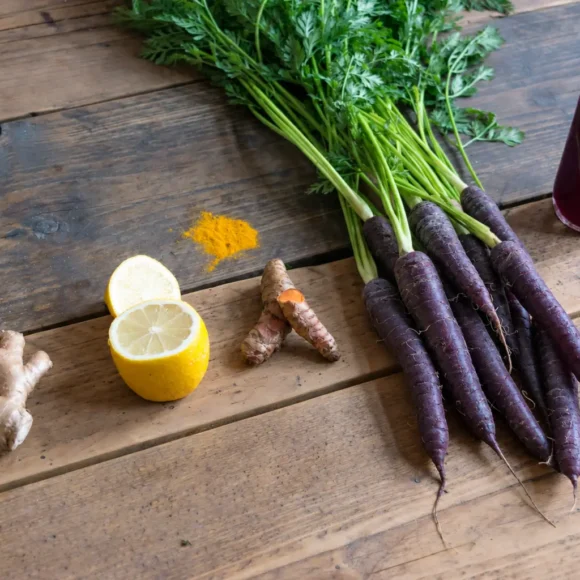 Black carrots with leafy green tops beside a bottle of purple juice, lemon, ginger and turmeric on a wooden table.