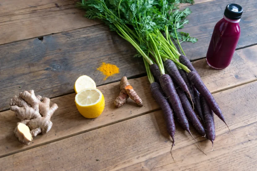Black Carrot Juice: Easy Homemade Recipe and Benefits Black carrots with leafy green tops beside a bottle of purple juice, lemon, ginger and turmeric on a wooden table.