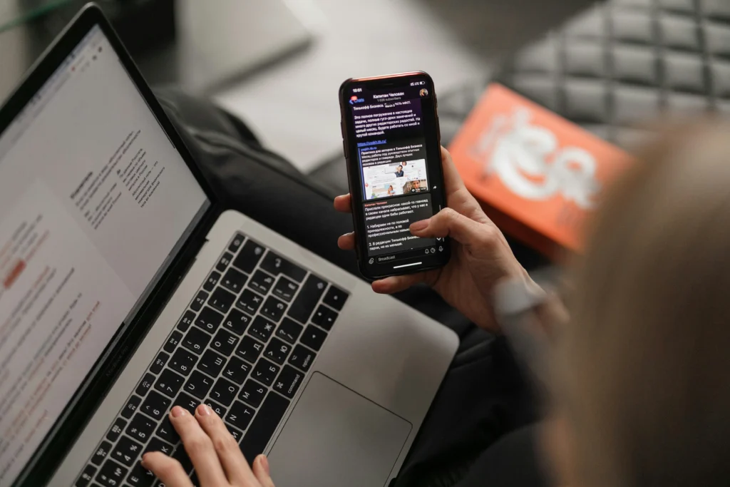 Person checking news updates on a smartphone while using a laptop, illustrating how to stop doomscrolling can interrupt daily activities and screen time habits.