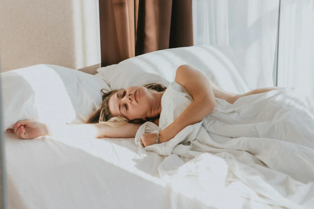 Woman relaxing in bed under soft white blankets, enjoying a peaceful Sunday morning as part of her Sunday reset routine.
