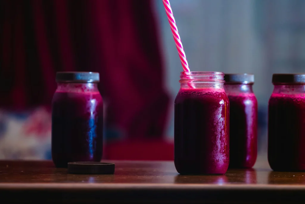 Black carrot juice shot in a glass jar with a pink striped straw, alongside three jars of deep purple juice on a wooden table.